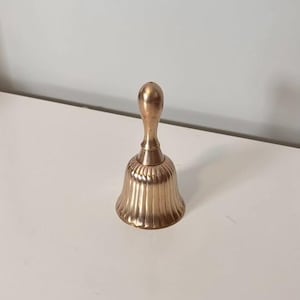 Pode incluir: Uma campainha de m&atilde;o em metal dourado com um cabo liso e arredondado, e um corpo alargado e canelado com sulcos verticais. A campainha tem um acabamento polido que reflete a luz, criando reflexos subtis ao longo da sua estrutura curva.