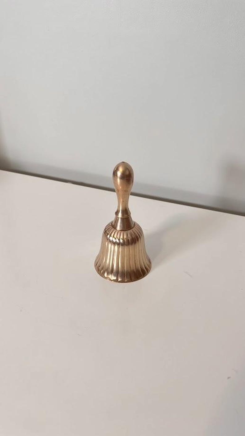 Pode incluir: Uma campainha de m&atilde;o em metal dourado com um cabo liso e arredondado, e um corpo alargado e canelado com sulcos verticais. A campainha tem um acabamento polido que reflete a luz, criando reflexos subtis ao longo da sua estrutura curva.