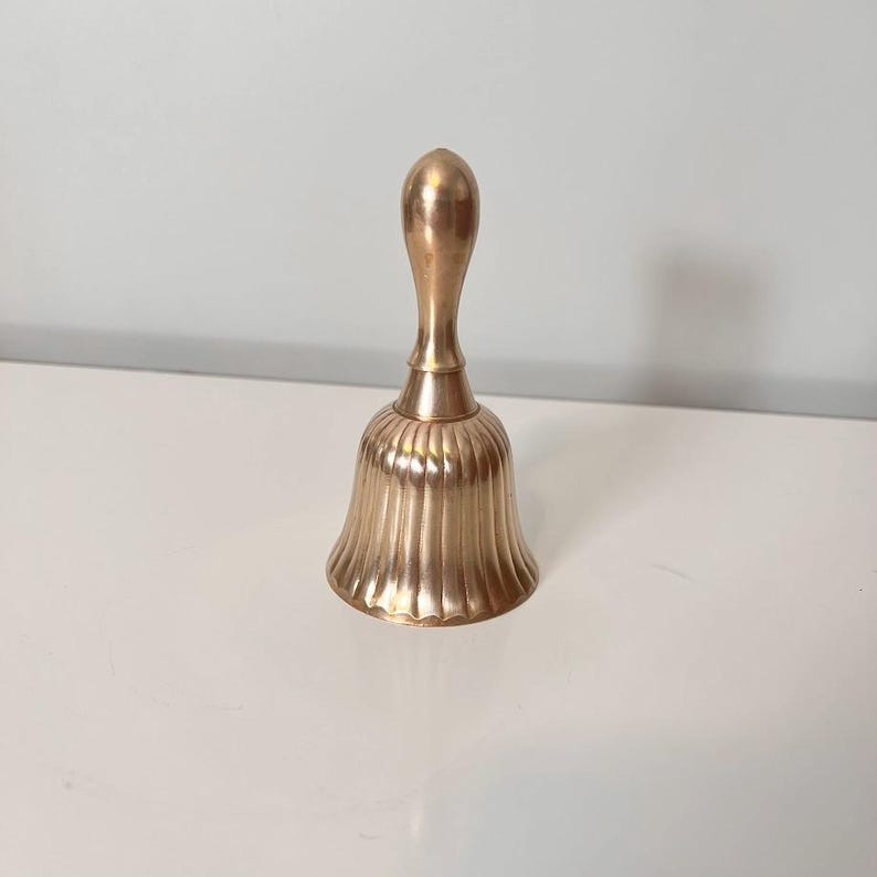 Pode incluir: Uma campainha de m&atilde;o em lat&atilde;o polido com um cabo liso e alongado e um corpo alargado e estriado com cristas verticais. A superf&iacute;cie met&aacute;lica tem um tom dourado quente e reflexivo, apresentando um design cl&aacute;ssico com topo arredondado.