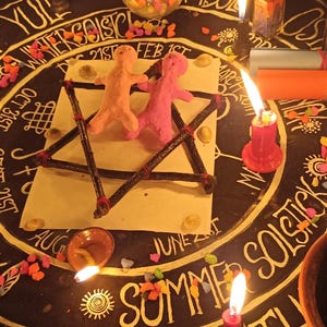 Puede incluir: Un mantel de altar circular negro con un dise&ntilde;o blanco de la rueda del a&ntilde;o que incluye texto como Samhain, Yule y Summer Solstice. Dos peque&ntilde;as figuras de arcilla se encuentran dentro de un pentagrama de ramas sobre una tarjeta cuadrada blanca.