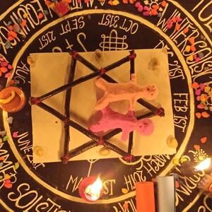 K&ouml;nnte beinhalten: Eine dunkle Oberfl&auml;che mit einem kreisf&ouml;rmigen Design mit Text und Daten, umgeben von kleinen bunten Steinen. Ein zentrales Quadrat enth&auml;lt eine sternartige Struktur mit zwei Figuren. Angez&uuml;ndete Kerzen und gerollte Papierr&ouml;hren sind ebenfalls vorhanden.