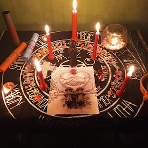 Puede incluir: Un altar ritual con una tela negra con un dise&ntilde;o circular blanco y texto. Se encienden velas rojas alrededor del altar, con una peque&ntilde;a estatua y otros objetos. Tambi&eacute;n hay piedras de colores y un candelabro de cristal.