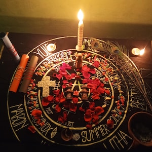 Puede incluir: Un altar iluminado con velas con una tela negra con un dise&ntilde;o circular con texto y s&iacute;mbolos. El altar incluye una vela encendida, p&eacute;talos de rosa rojos, una cruz y peque&ntilde;os objetos decorativos. Tambi&eacute;n hay una pluma de pavo real y pergaminos enrollados.