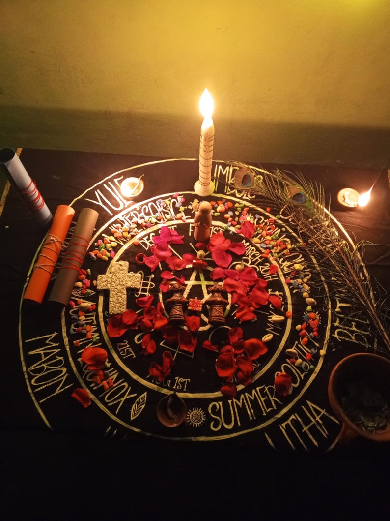 Puede incluir: Un altar iluminado con velas con una tela negra con un dise&ntilde;o circular con texto y s&iacute;mbolos. El altar incluye una vela encendida, p&eacute;talos de rosa rojos, una cruz y peque&ntilde;os objetos decorativos. Tambi&eacute;n hay una pluma de pavo real y pergaminos enrollados.