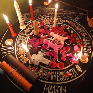 Puede incluir: Un mantel de altar circular negro con letras y s&iacute;mbolos blancos, rodeado de velas encendidas de varios colores y tama&ntilde;os. El altar presenta p&eacute;talos de rosa rosas, piedras de colores y dos figuras de mu&ntilde;ecas rosas. Tambi&eacute;n hay dos rollos enrollados y una cruz decorativa.