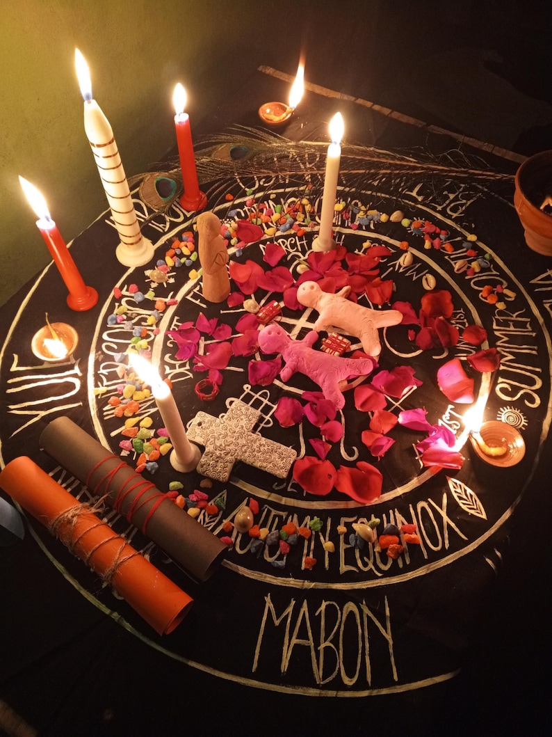 Puede incluir: Un mantel de altar circular negro con letras y s&iacute;mbolos blancos, rodeado de velas encendidas de varios colores y tama&ntilde;os. El altar presenta p&eacute;talos de rosa rosas, piedras de colores y dos figuras de mu&ntilde;ecas rosas. Tambi&eacute;n hay dos rollos enrollados y una cruz decorativa.