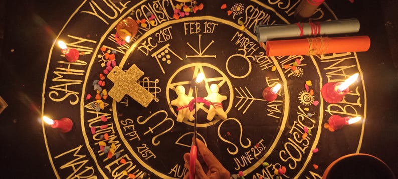 May include: A black circular altar cloth featuring gold printed text and symbols representing the wheel of the year, including dates like Dec 21st and Feb 1st. Two small white figurines, a textured cross, lit candles, and two rolled scrolls are placed on top.