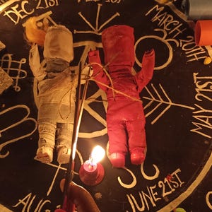 May include: Two fabric dolls, one beige and one red, are positioned on a circular black cloth featuring white text and symbols representing dates like Dec 21st, March 20th, May 21st, and June 21st. A textured cross, lit candles, and rolled scrolls are also present.