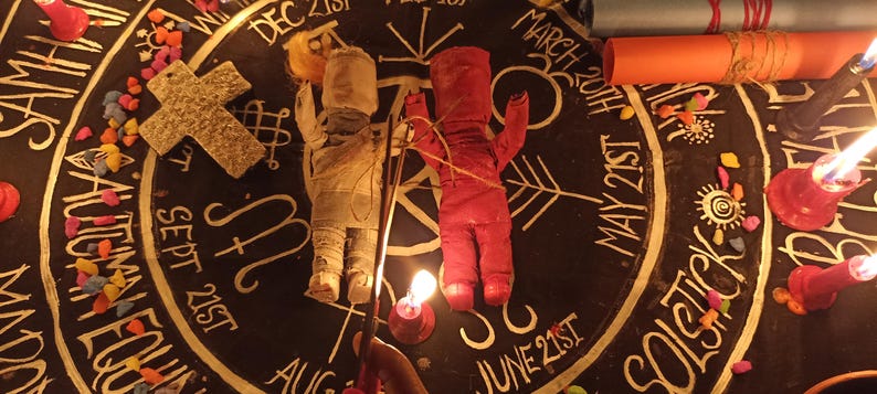 May include: Two fabric dolls, one beige and one red, are positioned on a circular black cloth featuring white text and symbols representing dates like Dec 21st, March 20th, May 21st, and June 21st. A textured cross, lit candles, and rolled scrolls are also present.
