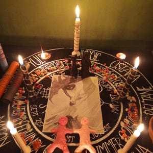 Puede incluir: Una escena ritual&iacute;stica con una tela negra con inscripciones y s&iacute;mbolos blancos, rodeada de velas encendidas. Dos peque&ntilde;as figuras de arcilla y un dibujo son centrales. Piedras de colores y velas adicionales est&aacute;n dispuestas.