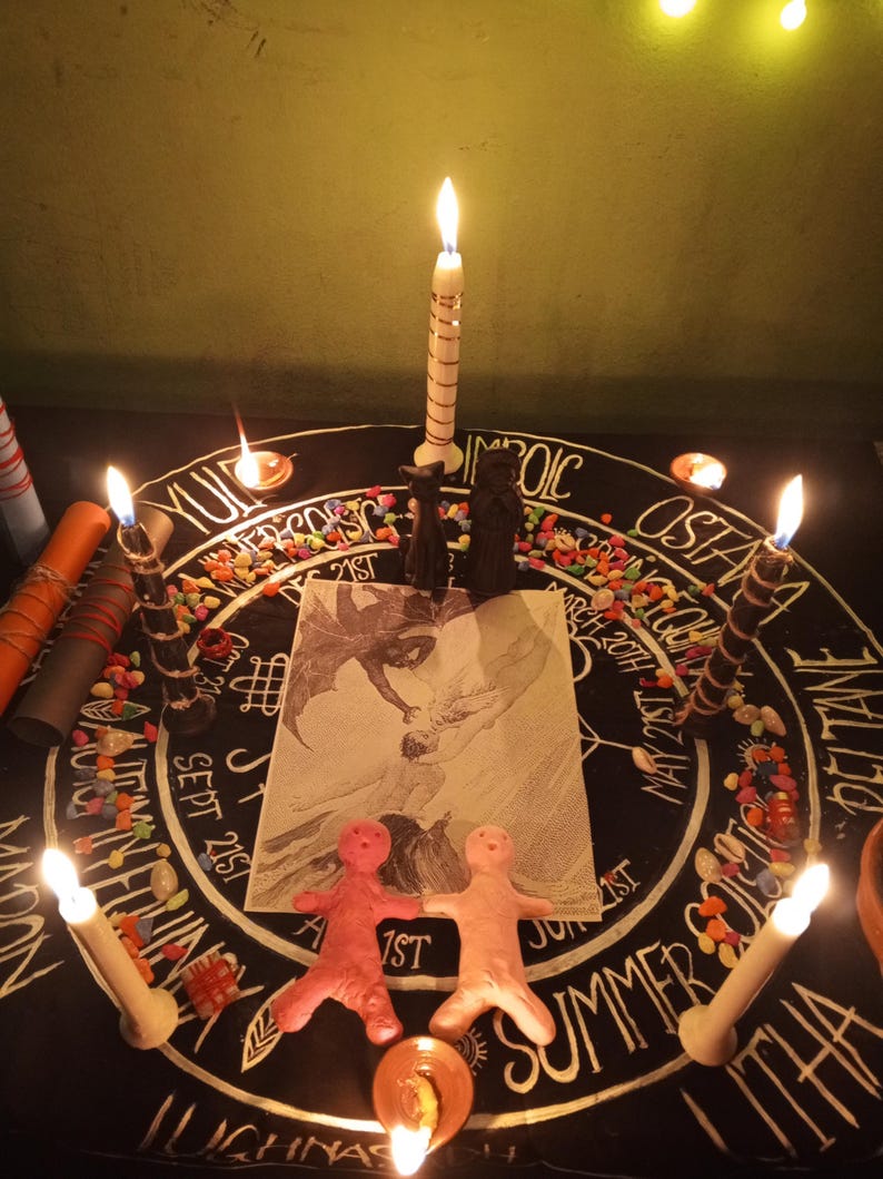 Puede incluir: Una escena ritual&iacute;stica con una tela negra con inscripciones y s&iacute;mbolos blancos, rodeada de velas encendidas. Dos peque&ntilde;as figuras de arcilla y un dibujo son centrales. Piedras de colores y velas adicionales est&aacute;n dispuestas.