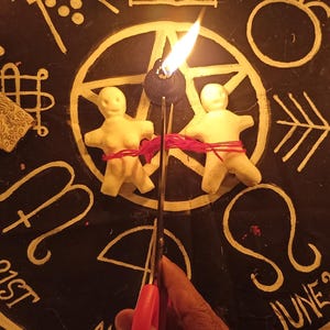 May include: Two off-white clay figures bound together with red string, positioned on a black circular cloth featuring gold astrological symbols, text including Sept 21st and June 21st, a textured cross, scattered colourful stones, and two lit candles.