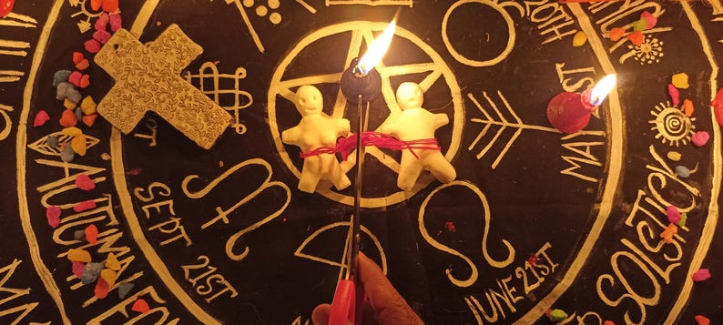May include: Two off-white clay figures bound together with red string, positioned on a black circular cloth featuring gold astrological symbols, text including Sept 21st and June 21st, a textured cross, scattered colourful stones, and two lit candles.