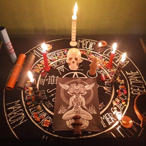 Puede incluir: Un altar ritual con una tela negra con texto y s&iacute;mbolos blancos. Velas encendidas, una calavera y una figura est&aacute;n dispuestas alrededor de una imagen central de una figura con alas. Tambi&eacute;n hay piedras de colores y papeles enrollados.