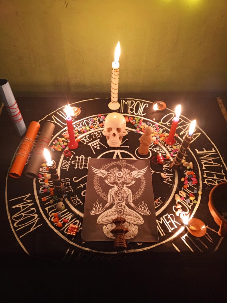 Puede incluir: Un altar ritual con una tela negra con texto y s&iacute;mbolos blancos. Velas encendidas, una calavera y una figura est&aacute;n dispuestas alrededor de una imagen central de una figura con alas. Tambi&eacute;n hay piedras de colores y papeles enrollados.