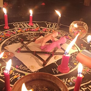 Puede incluir: Un pa&ntilde;o circular negro con una rueda del a&ntilde;o impresa en blanco que incluye textos como Samhain, Yul y fechas. Un marco de estrella de madera sostiene dos peque&ntilde;as figuras de arcilla, rodeadas por seis velas rojas encendidas y un cuenco.