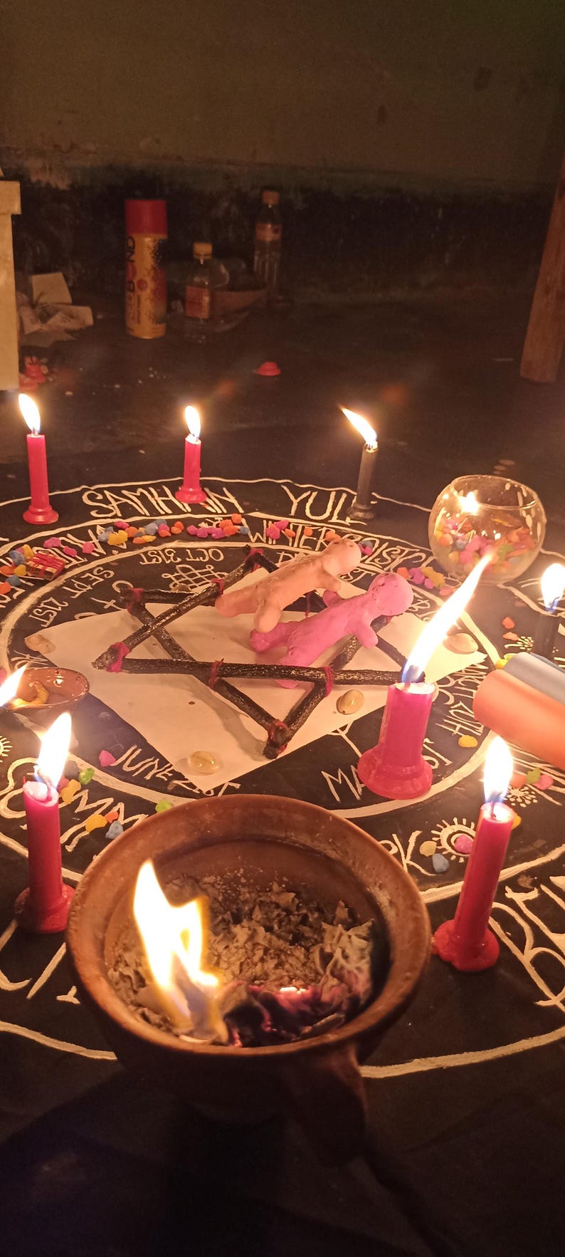 Puede incluir: Un pa&ntilde;o circular negro con una rueda del a&ntilde;o impresa en blanco que incluye textos como Samhain, Yul y fechas. Un marco de estrella de madera sostiene dos peque&ntilde;as figuras de arcilla, rodeadas por seis velas rojas encendidas y un cuenco.