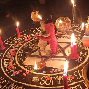 Puede incluir: Una alfombra circular negra con texto blanco de meses y eventos estacionales, dispuesta con m&uacute;ltiples velas encendidas, un peque&ntilde;o cuenco de cristal, dos pergaminos de papel enrollados y dos peque&ntilde;as figuras de tela en el centro de un pentagrama.