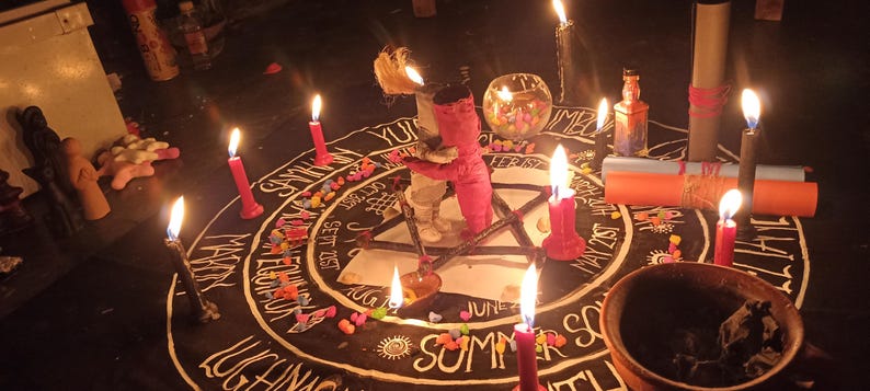 Puede incluir: Una alfombra circular negra con texto blanco de meses y eventos estacionales, dispuesta con m&uacute;ltiples velas encendidas, un peque&ntilde;o cuenco de cristal, dos pergaminos de papel enrollados y dos peque&ntilde;as figuras de tela en el centro de un pentagrama.