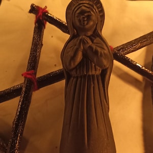 K&ouml;nnte beinhalten: Eine braune Statue einer Frau in einem langen Kleid mit gefalteten H&auml;nden im Gebet. Die Statue befindet sich vor einer sternf&ouml;rmigen Struktur aus dunkelbraunen St&ouml;cken, die mit roter Schnur zusammengebunden sind. Ein Glasbeh&auml;lter mit bunten S&uuml;&szlig;igkeiten befindet sich im Hintergrund.