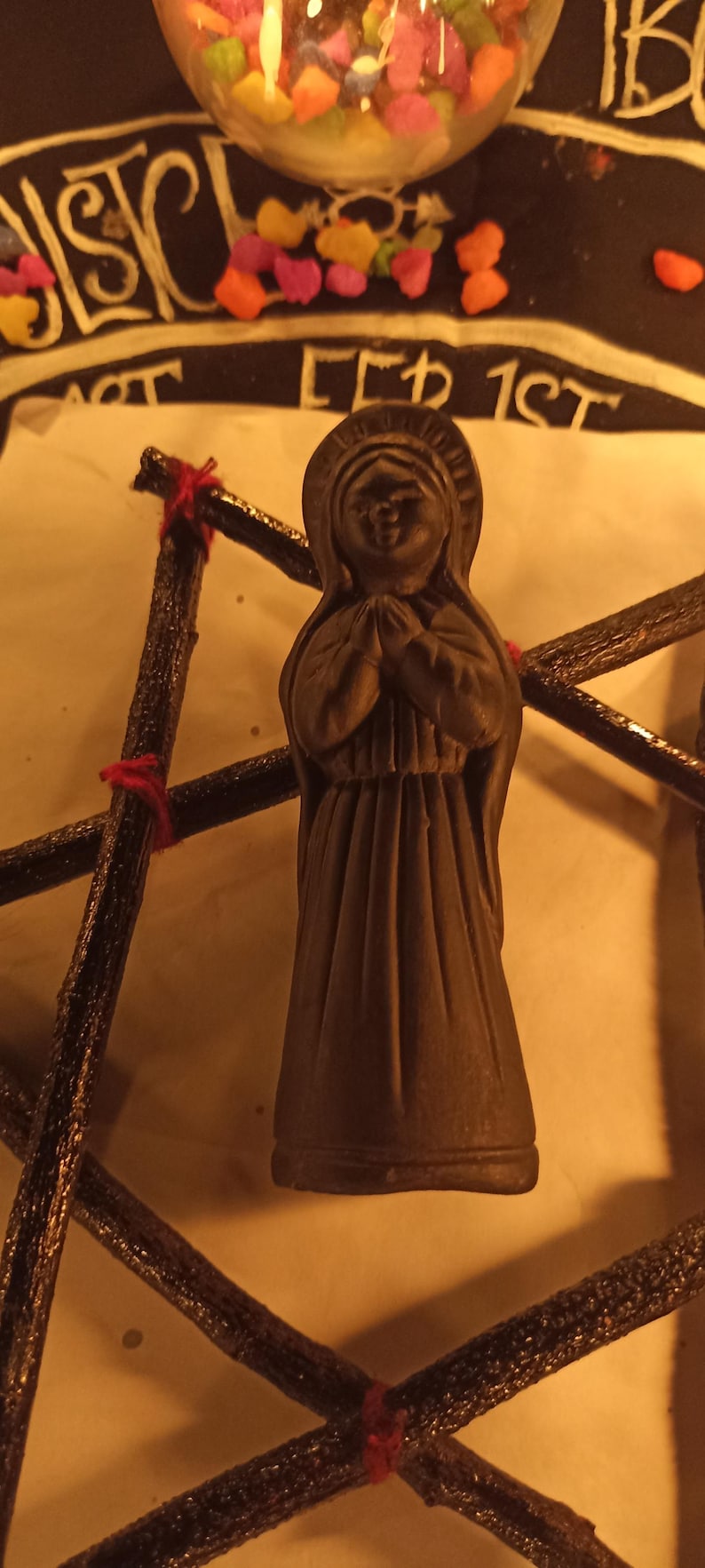 K&ouml;nnte beinhalten: Eine braune Statue einer Frau in einem langen Kleid mit gefalteten H&auml;nden im Gebet. Die Statue befindet sich vor einer sternf&ouml;rmigen Struktur aus dunkelbraunen St&ouml;cken, die mit roter Schnur zusammengebunden sind. Ein Glasbeh&auml;lter mit bunten S&uuml;&szlig;igkeiten befindet sich im Hintergrund.