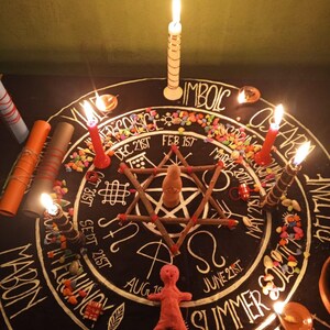 K&ouml;nnte beinhalten: Ein Ritualaltar mit schwarzer Oberfl&auml;che, mit einer sternf&ouml;rmigen Anordnung von St&ouml;cken und einer kleinen Puppe. Angez&uuml;ndete Kerzen umgeben den Altar, zusammen mit bunten Steinen und handschriftlichem Text. Die W&ouml;rter "MABON" und "SUMMER" sind sichtbar.