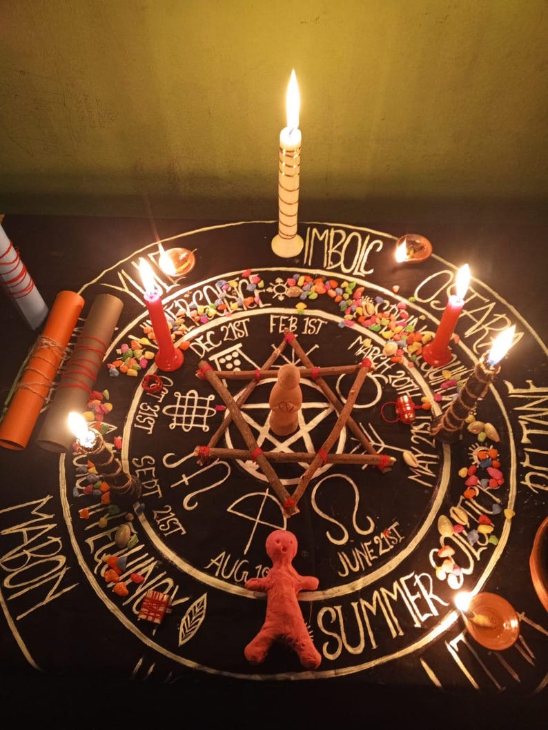 K&ouml;nnte beinhalten: Ein Ritualaltar mit schwarzer Oberfl&auml;che, mit einer sternf&ouml;rmigen Anordnung von St&ouml;cken und einer kleinen Puppe. Angez&uuml;ndete Kerzen umgeben den Altar, zusammen mit bunten Steinen und handschriftlichem Text. Die W&ouml;rter "MABON" und "SUMMER" sind sichtbar.