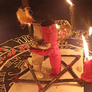 K&ouml;nnte beinhalten: Eine rituelle Szene mit einem zentralen Pentagramm, umgeben von brennenden roten Kerzen und einer puppenartigen Figur, die eine andere umarmt. Die Szene spielt sich auf einer dunklen Oberfl&auml;che mit handschriftlichem Text und bunten Steinen ab, was eine mystische Atmosph&auml;re schafft.