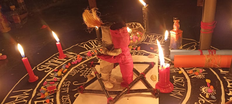 K&ouml;nnte beinhalten: Eine rituelle Szene mit einem zentralen Pentagramm, umgeben von brennenden roten Kerzen und einer puppenartigen Figur, die eine andere umarmt. Die Szene spielt sich auf einer dunklen Oberfl&auml;che mit handschriftlichem Text und bunten Steinen ab, was eine mystische Atmosph&auml;re schafft.