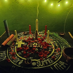 Puede incluir: Un mantel de altar negro con letras blancas y un dise&ntilde;o central, rodeado de velas, p&eacute;talos de rosa y peque&ntilde;os objetos. Una vela central est&aacute; encendida, con humo ascendente. La escena est&aacute; iluminada por luces de cadena.