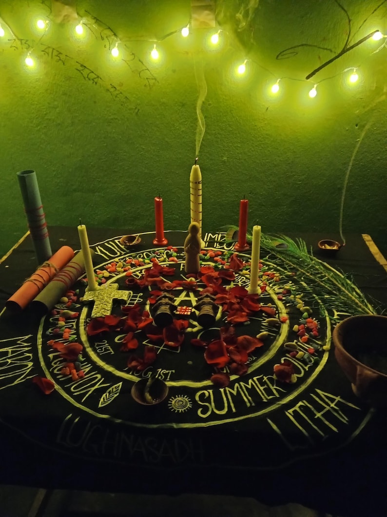 Puede incluir: Un mantel de altar negro con letras blancas y un dise&ntilde;o central, rodeado de velas, p&eacute;talos de rosa y peque&ntilde;os objetos. Una vela central est&aacute; encendida, con humo ascendente. La escena est&aacute; iluminada por luces de cadena.
