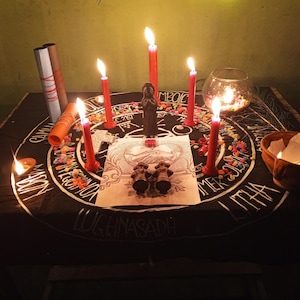 Puede incluir: Un altar ritual con una tela negra con letras blancas y un dise&ntilde;o circular. Velas rojas encendidas, una peque&ntilde;a estatua y un cuenco de vidrio con velas est&aacute;n dispuestos en el altar. Tambi&eacute;n hay un peque&ntilde;o cuenco de arcilla con una llama.