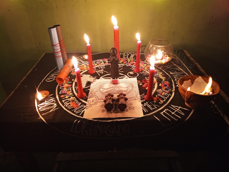 Puede incluir: Un altar ritual con una tela negra con letras blancas y un dise&ntilde;o circular. Velas rojas encendidas, una peque&ntilde;a estatua y un cuenco de vidrio con velas est&aacute;n dispuestos en el altar. Tambi&eacute;n hay un peque&ntilde;o cuenco de arcilla con una llama.
