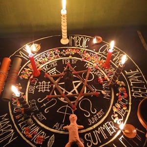 Puede incluir: Un altar circular negro con letras y s&iacute;mbolos blancos, rodeado de velas encendidas. En el centro hay una disposici&oacute;n de estrella y palos, con una peque&ntilde;a figura de arcilla en la parte inferior. Se ven piedras de colores y el texto "IMBOLC".