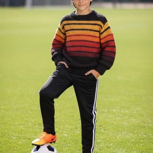 May include: A person stands on a football on a green pitch, wearing a striped jumper in shades of orange, yellow, and black. They are wearing black track pants with white stripes and orange football boots.