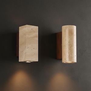 Può includere: Due applique da parete con finitura in pietra beige chiaro. Uno è rettangolare, l'altro cilindrico. Entrambi sono montati su un supporto in legno scuro ed emettono un bagliore caldo. Lo sfondo è grigio scuro.