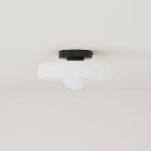 May include: A contemporary flush mount ceiling light. It has a black circular base and a white, rounded glass shade. The light bulb is visible through the bottom of the shade. The fixture is mounted on a white ceiling.