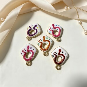 May include: Five white enamel charms with gold-toned metal edges, each featuring a coiled snake in a different colour—purple, pink, orange, red, and dark red—surrounded by small colourful star accents and a circular loop at the bottom for attachment.