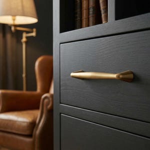 Könnte beinhalten: Nahaufnahme eines schwarzen Holzschranks mit einem goldfarbenen Griff. Der Griff ist horizontal und hat ein einzigartiges, geometrisches Design. Der Schrank ist Teil eines größeren Möbelstücks, möglicherweise eines Bücherregals oder einer Kommode. Im Hintergrund ein Ledersessel und eine Lampe.