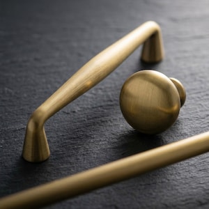 以下が含まれることがあります： 真鍮製のキャビネット金具のクローズアップ。長いプルハンドルと丸いノブが含まれています。 金具はブラッシュ仕上げで、暖かく金色です。アイテムは、暗く質感のある背景に配置されています。