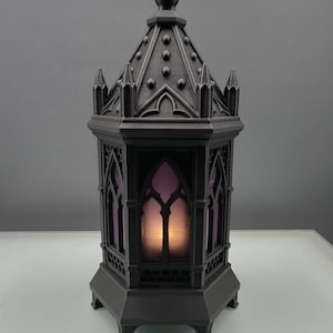 May include: A matte black lantern with a hexagonal structure and pointed gothic arches. The frame features decorative studs on the roof and purple translucent panels. A warm, glowing light is visible from within the center of the lantern.