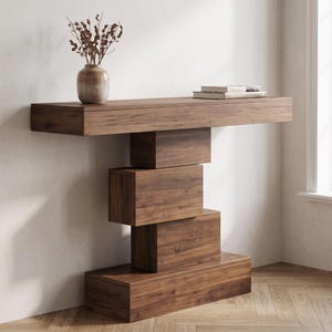 May include: A dark brown wooden console table featuring a long rectangular top supported by a vertical stack of four offset rectangular wooden blocks. A ceramic vase with dried branches and two stacked books sit on the table surface.