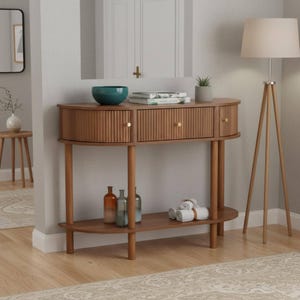May include: A mid-century modern style console table in light brown wood. The table has a half-moon shape with two drawers and a lower shelf. Decorative items include a teal bowl, books, a small plant, and bottles.