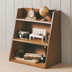 Puede incluir: Una estantería de madera de tres niveles, que muestra objetos decorativos. La balda superior contiene un reloj dorado y un globo terráqueo. La balda central presenta una cámara en miniatura y un banco blanco. La balda inferior tiene una piña, un calendario de madera y una figura de gato negro.