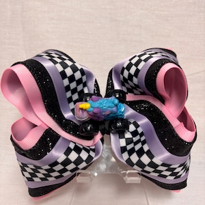 May include: A large hair bow featuring layered ribbons in black glitter, light purple, and a black and white checkered pattern. A small blue and purple toy monster truck with a yellow horn is attached to the center of the bow as an embellishment.