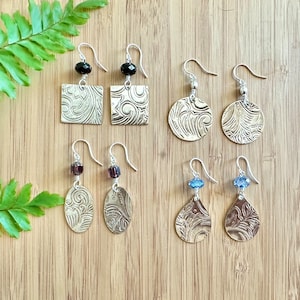 May include: Six pairs of silver earrings with different colored beads and patterned metal charms. The charms are square, round, and oval shaped. The earrings are displayed on a wooden surface.