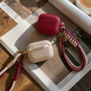 May include: Two wireless earbud cases are shown, one deep red, the other light beige. Each case has a lanyard with a braided design in red and white. The lanyards have a small tag that says "Hand Made". The cases are on a printed surface.