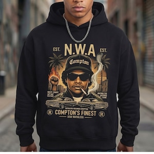 Puede incluir: Sudadera con capucha negra con un gráfico de un hombre con gafas de sol y una gorra de Compton, fumando un cigarrillo. El diseño incluye el texto "N.W.A", "Compton's Finest" y "Ruthless Records Compton". También se representan palmeras y coches clásicos.