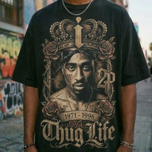 Puede incluir: Camiseta negra con un retrato de Tupac Shakur, corona, rosas y las palabras "Thug Life" y "1971-1996". El diseño utiliza tonos marrones, beige y dorados.
