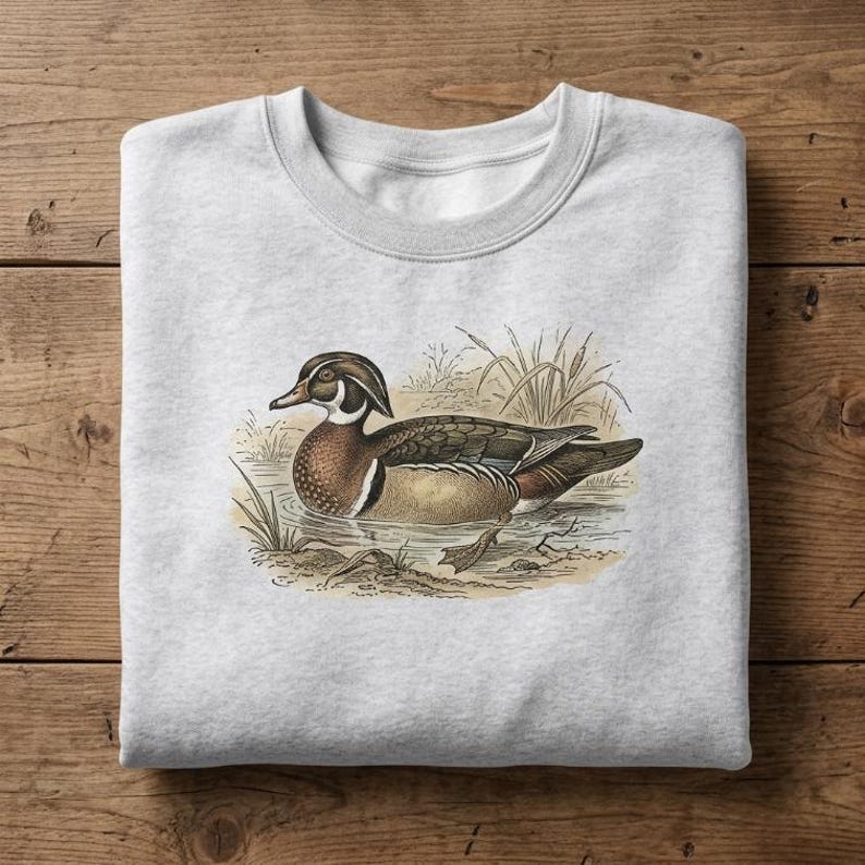 May include: A light gray crewneck sweatshirt featuring a detailed illustration of a wood duck swimming in water with reeds. The duck has brown, white, and iridescent green plumage. The graphic is centered on the front of the garment.